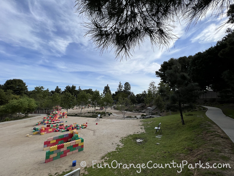 wide view of park with sidewalk on the right and dirt areas with colorful large lego like blocks for building amongst trees and under a blue sky