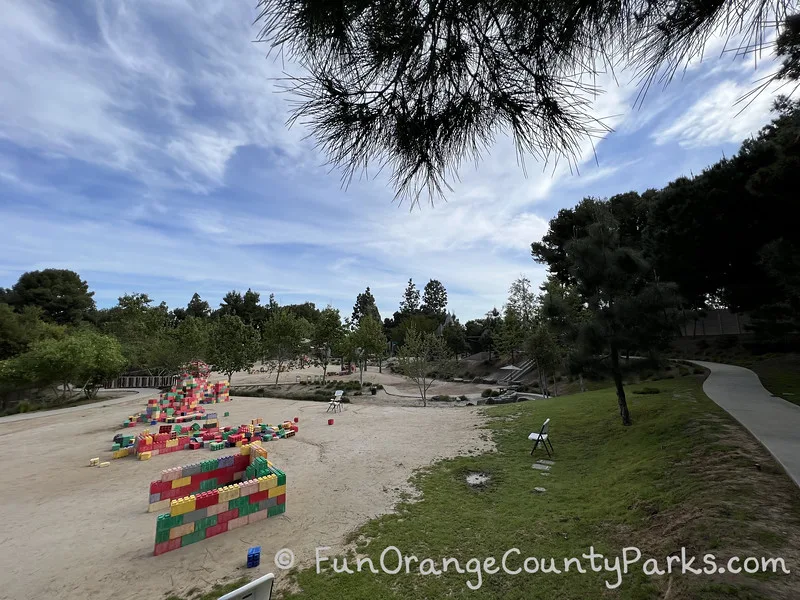 wide view of park with sidewalk on the right and dirt areas with colorful large lego like blocks for building amongst trees and under a blue sky