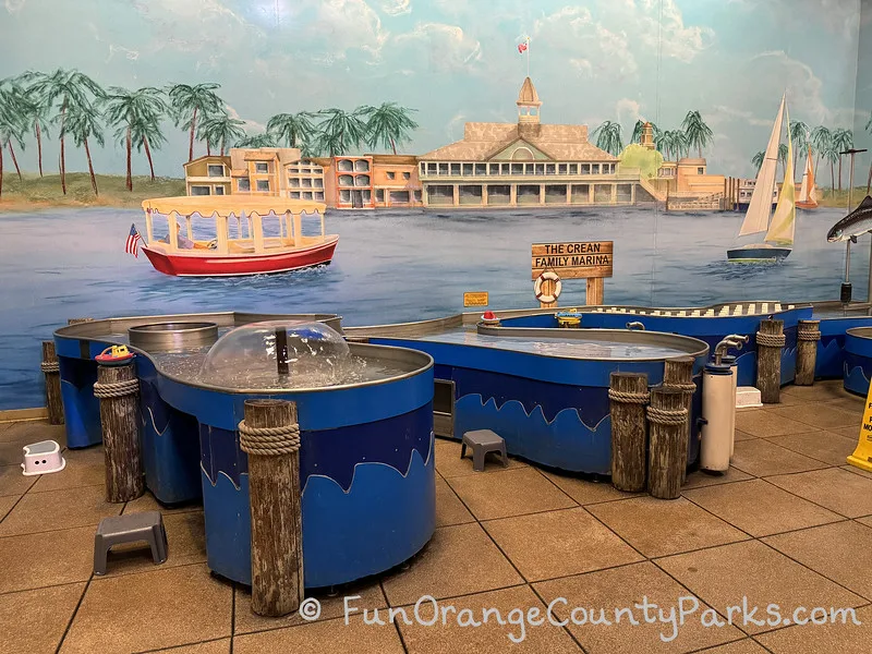 a mural depicting balboa peninsula pavilion building with an electric boat and sailboat in the harbor behind blue pools and fountains for children to play with floating objects at child level